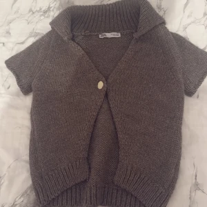 Brun stickad kofta från Zara - Kortärmad brun stickad kofta från Zara med bred krage och en snygg guldfärgad knapp framtill. Koftan har ribbade muddar och en relaxed passform, perfekt att slänga över en t-shirt eller linne för en chill look.