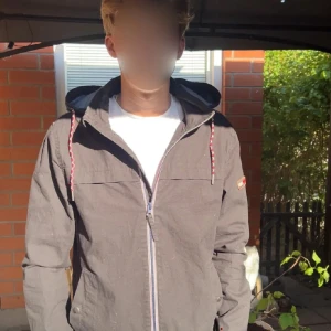 Svart vindjacka från Hilfiger Denim - Svart vindjacka från Hilfiger Denim med huva och snygga röda/vita snören. Jackan har dragkedja framtill och en enkel, clean design som passar perfekt till streetwear. Materialet är bomull och jackan har normal passform. Perfekt för vår och höst.