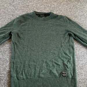 Mörkgrön stickad tröja från Replay - Snygg mörkgrön stickad tröja från Replay med rund hals och ribbade muddar vid ärmslut och nederkant. Tröjan har en diskret Replay-logga nedtill och är perfekt för dig som gillar stilrena och enkla plagg. Passar dig som vill ha en chill och klassisk look.