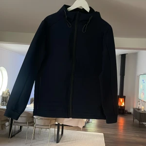 Mörkblå vindjacka med huva - Snygg mörkblå vindjacka med hel dragkedja och justerbar huva med snörning. Jackan har en clean look, raka linjer och långa ärmar. Perfekt för dig som gillar stilren och funktionell design. Fodrad insida för extra komfort.