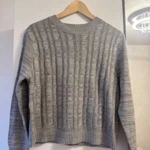 Grå kabelstickad tröja med rund hals - Snygg grå kabelstickad tröja med rund hals och ribbade muddar. Tröjan har ett klassiskt kabelmönster på framsidan och är enfärgad baktill. Använd 3-4 gånger.