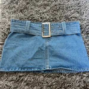 Blå jeanskjol med bälte från NLY Jeans - Säljer en snygg blå jeanskjol från NLY Jeans med bredt bälte och silverspänne i midjan. Kjolen är kort och har dragkedja i sidan för enkel påtagning. Perfekt för dig som gillar denim och vill ha en cool look.