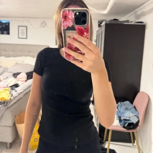 Svart tight t-shirt i bomull - En enkel svart t-shirt med korta ärmar och rund hals. Modellen är tight och sitter snyggt mot kroppen. Materialet känns mjukt och stretchigt, troligtvis bomull. Perfekt att matcha med jeans eller shorts för en clean look.