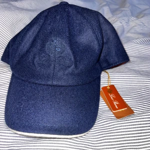 Mörkblå keps från Loro Piana - Exklusiv mörkblå keps från Loro Piana med broderad logga framtill. Utsidan är i mjuk ull och insidan har ett rutigt foder i bomull och orange kant. Justerbar baktill med metallspänne. Perfekt accessoar för en clean och stilren look. 0bs lnte 0rglnal!