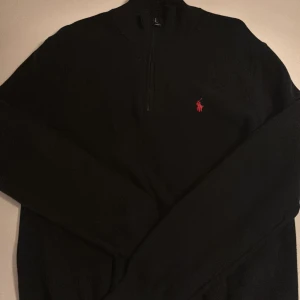 Svart half zip tröja Polo Ralph Lauren - Svart långärmad tröja från Polo Ralph Lauren i mjuk pima bomull. Klassisk half zip med hög krage och röd broderad logga på bröstet. Perfekt för dig som gillar stilren och tidlös design med en sportig touch. 