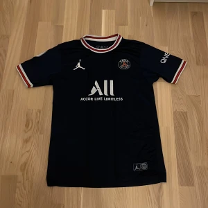 PSG x Jordan marinblå fotbollströja M - Snygg PSG fotbollströja i marinblått med vita och röda detaljer från Jordan. Kortärmad modell med broderat PSG-emblem och Jumpman-logga på bröstet. Rund hals med randig ribbad kant. Tillverkad i lätt och ventilerande polyester, perfekt för match eller träning.