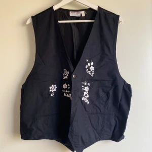 Svart väst med broderade detaljer - Svart väst från Berto Lucci med vit broderad design framtill. Västen har v-ringad hals, knappstängning och två fickor. Materialet känns som bomull och polyester, och broderierna ger en unik och cool vibe. Storleken är okänd men känns som M!