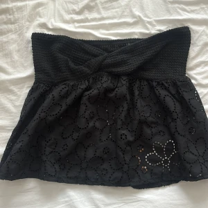 Svart topp med broderade blommor - ALDRIG ANVÄND! Supersöt svart topp med blommor i tyget. Toppen har en omlott-detalj upptill och är gjord i mjuk bomull. Perfekt för dig som gillar unika detaljer och vill ha en topp som sticker ut lite extra.