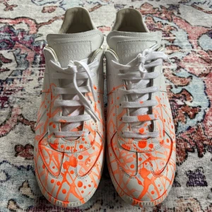 Färgstänkta sneakers från Maison Margiela - Unika sneakers från Maison Margiela med ett konstnärligt färgstänk orange på en vit bas. Skorna har snörning och en klassisk siluett som ger en cool och kreativ look.