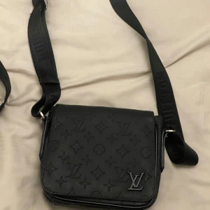 Svart axelväska - Snygg svart axelväska  i skinn med klassiskt monogrammönster. Väskan har justerbar axelrem med , ett stort fack och dragkedja på baksidan. Perfekt för dig som vill ha en stilren och lyxig accessoar.