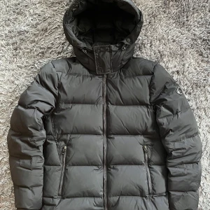 Svart pufferjacka från J.Lindeberg - Säljer en svart pufferjacka från J.Lindeberg med huva och dragkedja framtill. Jackan har två snedställda fickor med dragkedja och en snygg logga på ärmen. Perfekt för kalla dagar och riktigt stilren i sin design.