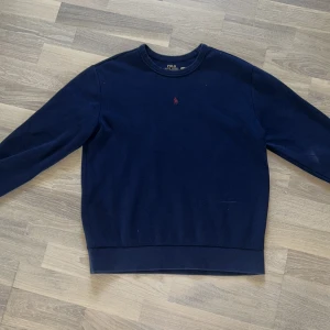 Mörkblå sweatshirt från Polo Ralph Lauren - Snygg mörkblå sweatshirt från Polo Ralph Lauren med klassisk röd broderad logga på bröstet. Tröjan har rund hals, långa ärmar och är gjord i mjuk bomull. Perfekt för dig som gillar stilrena och tidlösa plagg.  Nästan helt oanvänd