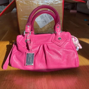 Vintage Rosa handväska Marc by Marc Jacobs - Säljer en rosa handväska från Marc by Marc Jacobs i skinn med silvriga detaljer och dubbla handtag. Väskan har även en avtagbar rosa Hello Kitty-berlock som jag själv matchat till väskan. Axelrem ingår. Mått ca 30 × 20 × 10 cm. Innerfodret är rent, metallen utan repor och väskan är i mycket gott skick