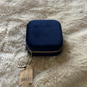 Mörkblå smyckesask med spegel - Snygg och praktisk mörkblå smyckesask med mjuk yta och guldfärgad dragkedja. Insidan har flera fack för ringar, örhängen och halsband samt en liten spegel i locket. Perfekt för att hålla dina favoritsmycken organiserade.