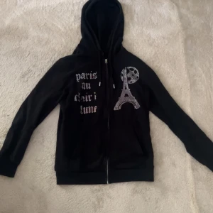 Svart hoodie med Paris-tryck och strass - Svart hoodie med dragkedja och huva, dekorerad med strassmotiv av Eiffeltornet och måne samt texten 'paris au clair lune' på framsidan. Långärmad och med snörning i huvan. Perfekt för dig som gillar coola detaljer och Paris-vibes.