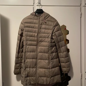 Beige lång pufferjacka med huva - Säljer en beige pufferjacka med huva och dragkedja framtill. Jackan är lång, quiltad och har en clean look utan synliga loggor. Perfekt för kyliga dagar och enkel att styla till olika outfits. Långärmad och med praktisk passform.