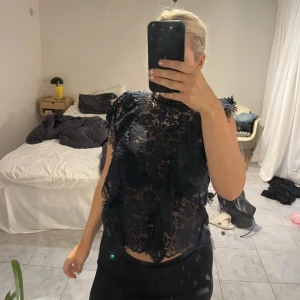 Mörkblå spetsblus med volangdetaljer - Säljer en svart blus i genombruten spets med blommigt mönster och volangdetaljer längs axlar och framstycke. Blusen är ärmlös och har en rak passform, Stängs med knapp där bak. 