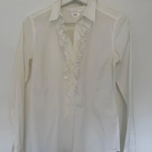 Vit volangskjorta från Filippa K - Snygg vit skjorta från Filippa K med volangdetaljer längs knäppningen framtill. Klassisk krage och långa ärmar. Skjortan är gjord i bomull och har en stilren, tidlös look som passar perfekt till jeans eller kostymbyxor. Aldrig använd🌟