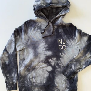 Svart och grå tie-dye hoodie Nudie Jeans - Säljer en hoodie från Nudie Jeans Co i storlek M. Tröjan har ett snyggt svart och grått tie-dye mönster, huva med snörning och stor magficka. Tryck med NJ CO på bröstet. Tillverkad i 100% ekologisk bomull, perfekt för chill dagar.