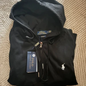 Svart hoodie från Polo Ralph Lauren - Svart hoodie från Polo Ralph Lauren med dragkedja och huva. Helt ny med prislappen kvar. Custom fit. Storlek M. Nypris: ~1700 | Vi säljer för 999. Tveka inte att höra av er med prisförslag eller funderingar!