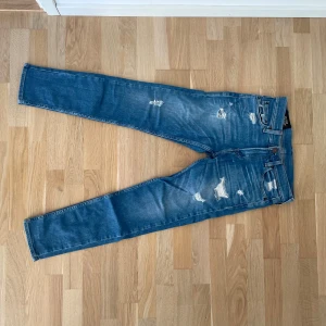 Slitage jeans  - Säljer ett par blå jeans från Hollister med slitna detaljer och snygga rippade partier. Jeansen har klassisk femficksdesign, normal midja och är tillverkade i stretchigt denim för en tajt passform. Perfekt för dig som gillar en trendig och avslappnad stil.
