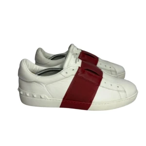Valentino Open sneakers vit/röd  - Snygga Valentino Open sneakers i vitt skinn med bred röd läderdetalj över vristen. Klassisk rund tå och platt sula med ikoniska nitar baktill. Perfekta för dig som vill sticka ut med en lyxig och stilren look. BARA ANVÄNDA 2 GÅNGER!!!
