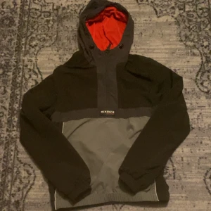 McKenzie windbreaker  - Snabb leverans! I fantastisk skick! Precis om ny!Snygg vindjacka från McKenzie i svart och grått med röd meshfoder. Jackan har huva med snörning, reflekterande detaljer och elastiska muddar. Perfekt för blåsiga dagar och har en sportig vibe. Lätt och smidig att bära, med dragkedja framtill och logga på bröstet.