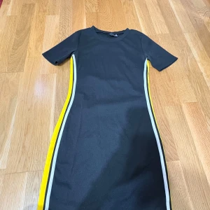 Svart kortklänning med gula ränder - Snygg svart kortklänning med korta ärmar och coola gula och vita ränder längs sidorna. Klänningen har en enkel, figurnära passform och rund halsringning. Perfekt för dig som gillar sportig och modern stil.