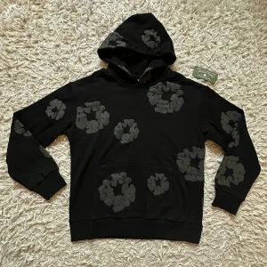 DENIM TEARS HOODIE- svart med lapparna kvar - Svart hoodie från Denim Tears med gråa blommönster över hela tröjan, inklusive huvan. Tröjan har en klassisk känguruficka och ribbade muddar. Perfekt för dig som gillar streetwear och vill sticka ut med en unik design. Helt ny och oanvänd och passar helt perfekt till dig som vill ha en lite mer unik stil fast fortfarande falla inom street/ grisch stilen! 