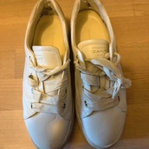 Vita sneakers från Gant i skinn - Snygga vita sneakers från Gant med breda tygsnören och guldfärgade öljetter. Skorna är i skinn och har en klassisk rund tå och platt sula. Perfekta för en clean och stilren look. Kolla insidan av skon för defekt 