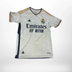 Real Madrid Bellingham #5 matchtröja - Säljer en officiell Real Madrid fotbollströja från Adidas med Bellingham och nummer 5 på ryggen. Tröjan är vit med marinblå detaljer och guldiga ränder på axlarna. V-ringad hals, korta ärmar och tryckta loggor. Materialet är lätt och andas, perfekt för match eller träning.