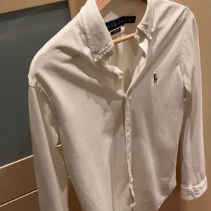 Vit skjorta från Ralph Lauren - Klassisk vit skjorta från Ralph Lauren med broderad logga på bröstet. Skjortan har lång ärm, knappar hela vägen och button down-krage. Tillverkad i mjuk bomull som känns skön mot huden. Perfekt för en clean och stilren look.