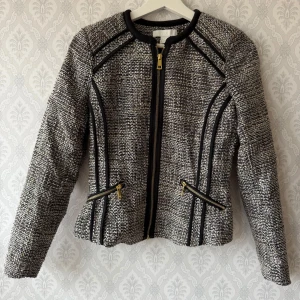 Grå kavajjacka med dragkedjor - Snygg gråmelerad kavajjacka från H&M med svarta detaljer och guldiga dragkedjor. Jackan har lång ärm, markerade sömmar och två snedställda fickor framtill. Materialet är strukturerat och ger en exklusiv känsla. Perfekt för dig som gillar stilrena och trendiga plagg.