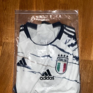 Italien adidas fotbollströja vit/blå S - Säljer en snygg Italien fotbollströja från adidas i storlek S. Tröjan är vit med marinblå mönster och detaljer, har korta ärmar och klassiska adidas-ränder på axlarna. Broderad Italien-logga på bröstet och tillverkad i ventilerande HEAT.RDY-material. 