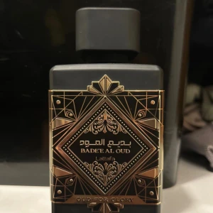 Lattafa Badee Al Oud Oud For Glory - Exklusiv parfymflaska i mattsvart med detaljer i guld och geometriskt mönster. Flaskan är fyrkantig med en robust svart kork och guldplakett på toppen. Doften är Eau de Parfum och rymmer 100 ml. Perfekt för dig som gillar orientaliska och lyxiga dofter.