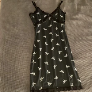 Svart kort klänning med fjärilar H&M - Snygg svart kort klänning från H&M DIVIDED i storlek XS. Klänningen har vita fjärilsmönster över hela tyget och tunna axelband. Spetsdetaljer vid halsringningen och nederkanten ger en extra touch. Perfekt för dig som gillar en cool och söt stil.