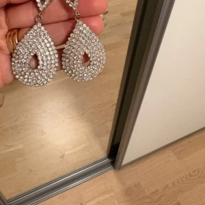  örhängen - Säljer ett par stora, droppformade örhängen i silverfärg med massor av glittrande stenar. De har en öppen mitt och är riktigt statement med sitt gnistrande utseende. Perfekta för att lyfta din look och ge extra bling. Använd 1 gång
