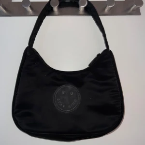 Svart axelväska  - Snygg svart axelväska från Stay Kind med broderad smiley och texten 'XO Stay Kind' på framsidan. Väskan har en mjuk, lätt glansig yta och stängs med dragkedja. Perfekt storlek för mobilen, plånboken och lite till.