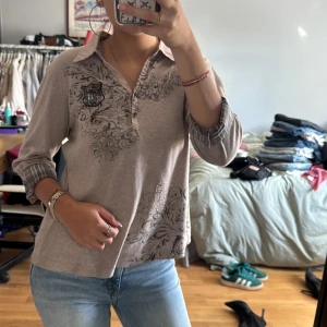 Beige pikétröja med mönster Gerry Weber - Snygg beige pikétröja från Gerry Weber med svart dekorativt mönster och emblem på bröstet. Krage och ärmslut har rutiga detaljer i grått och blått. Långärmad modell i mjukt bomullsmaterial. Stl står inte men gissar på runt m