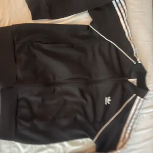 Svart Adidas track jacket med vita ränder - Säljer en klassisk svart Adidas track jacket med vita ränder längs ärmarna och broderad logga på bröstet. Jackan har dragkedja framtill, ståkrage och två fickor. Perfekt för en sportig och avslappnad stil.