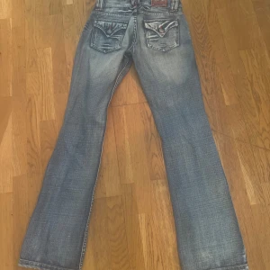 Kapora5 blå jeans med broderade fickor - Säljer ett par snygga blå jeans från Kaporal med coola broderade bakfickor och orangea detaljer. Jeansen är bootcut och storlek 37. Innerbenslängden är 81 och midjemåttet är 37 tvärs över 