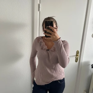 Ljusrosa Odd Molly ribbad topp - Supersöt ljusrosa ribbad topp från Odd Molly med lång ärm och spetsdetaljer vid v-ringad hals. Liten svart rosett framtill och vågig kant nedtill. 