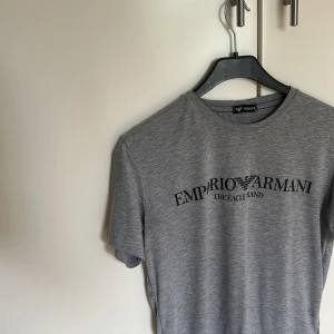 Grå Emporio Armani t-shirt - Snygg grå t-shirt från Emporio Armani med svart logotyptryck på bröstet. Klassisk rund hals och korta ärmar. Perfekt för dig som gillar stilrena och exklusiva märken. Den kommer i en neutral grå nyans som är lätt att matcha med både jeans, chinos eller shorts. Materialet känns mjukt och skönt mot huden. 