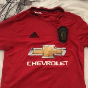 Manchester United röd fotbollströja Adidas - Säljer en röd Manchester United fotbollströja från Adidas med svart klubbmärke och Chevrolet-logga i guld och silver på bröstet. Tröjan har korta ärmar, rund hals med svart kant och är tillverkad i lätt, ventilerande material. Perfekt för match eller träning. Storlek 13-14 vilket motsvarar L fast i barnstorlek.