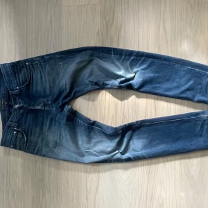 Blå jeans från Nudie Jeans - Storlek W29 W32                                            Snygga blå jeans från Nudie Jeans med slitna detaljer. Jeansen har en perfekt passform och slitningar framtill och baktill. Har en större slitning på höger sidan vid knäet. 