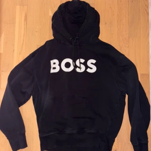 Svart hoodie från BOSS med tryck - Svart hoodie från BOSS med stor vit BOSS-logga på bröstet. Tröjan har huva med snörning, lång ärm och en klassisk känguruficka framtill. Materialet är mjuk bomull och passformen är normal. Perfekt för chill dagar eller när du vill ha en clean streetstyle.