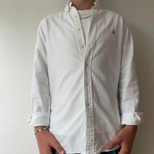 Vit skjorta från Ralph Lauren slim fit - Klassisk vit skjorta från Ralph Lauren i slim fit-modell. Skjortan har långa ärmar, knappar framtill och den ikoniska broderade loggan på bröstet. Tillverkad i bomull och passar perfekt till jeans eller chinos för en clean och stilren look.