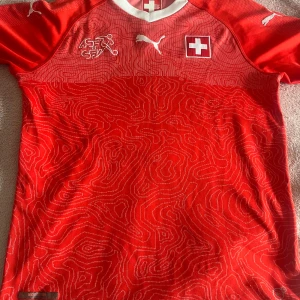 Röd Schweiz fotbollströja🇨🇭🧀 - Schweiz landslagströja från Puma i starkt röd färg med vita detaljer och Schweiz-emblem på bröstet. Tröjan har korta ärmar och ett unikt mönster som liknar topografiska linjer över hela framsidan. Materialet är lätt och andas, perfekt för match eller träning. Köpt i Schweiz 🇨🇭 