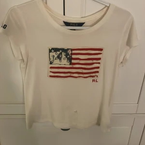 Vit t-shirt med flaga  Polo - Vit t-shirt från Polo Ralph Lauren med broderad amerikansk flagga framtill och RL-detalj. Flagan är sliten efter tvätt och ett hål ner till.