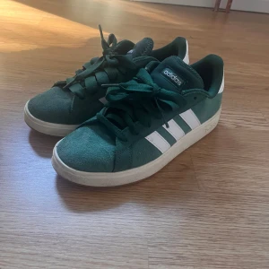 Gröna Adidas sneakers  - Snygga gröna Adidas sneakers med klassiska vita ränder och vit häl. Ovandel i mocka och platt sula. Skorna har snörning och en stilren, sportig vibe som passar perfekt till streetwear. Adidas-logga på plös och häl ger en clean look. Knappt använda alls, stel 40 men jag har 38/39 och upplever dom mer som 39. Ser ut som campus men är en liknande modell från adidas💗 skriv om ni har någon fråga. 